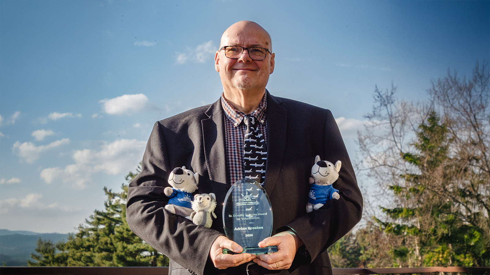 Adrian Brookes in a suit outside smiling and holding UHKF Volunteer Award and mascot Asher stuffies.