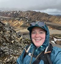 Megan Wheatley wearing a rain jacket and backpack, standing at the top of a mountain.