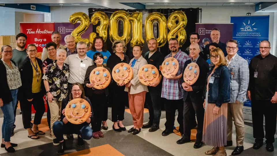 Smile Cookie Week funding announcement at Kingston General Hospital. Hospital and Tim Hortons staff holding smile cookies in front of number balloons.