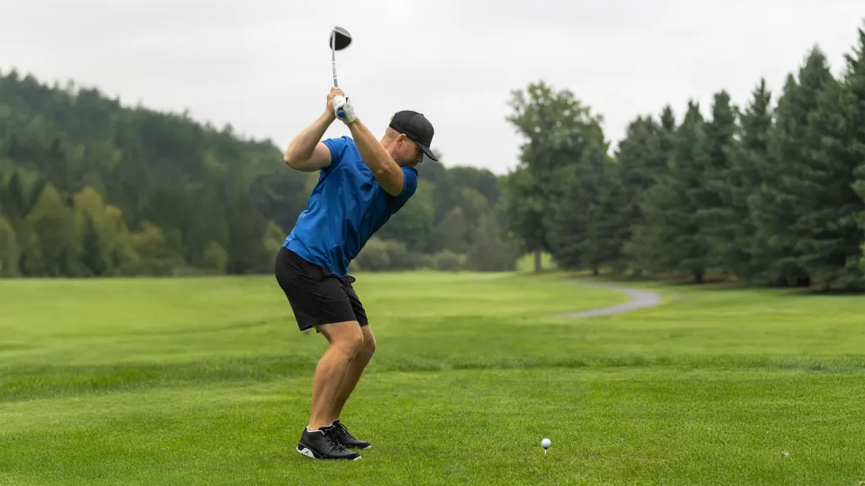 Man swinging golf club on the green in a blue golf shirt and black hat.
