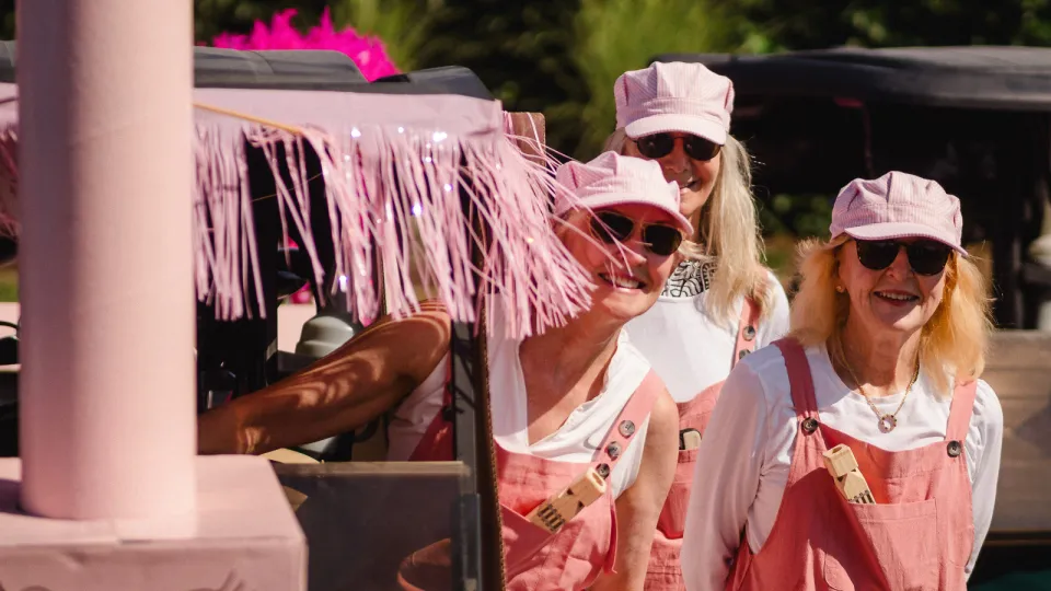 Golfers at the Rose of Hope Golf Tournament in pink golfing gear standing by their cart. Their outfits resemble train conductors, with pink hats and overalls.
