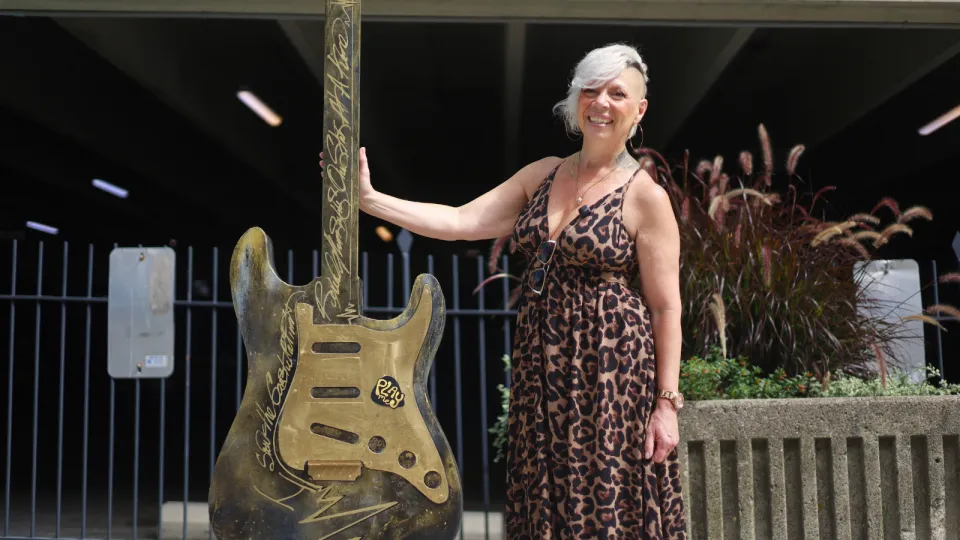 Cher Reynolds standing outside Hotel Dieu Hospital with her guitar art installation. The guitar stands at six feet tall and incorporates EGK lines in the design, weaving in the themes of healthcare to the Kingston story of music and art.