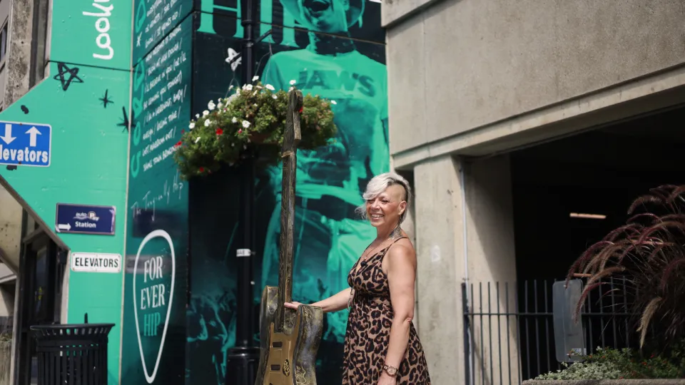 Cher Reynolds standing outside Hotel Dieu Hospital with her guitar art installation. The guitar stands at six feet tall and incorporates EGK lines in the design, weaving in the themes of healthcare to the Kingston story of music and art. In the background you see a wall wrap promoting The Tragically Hip's ten year anniversary since their last show.