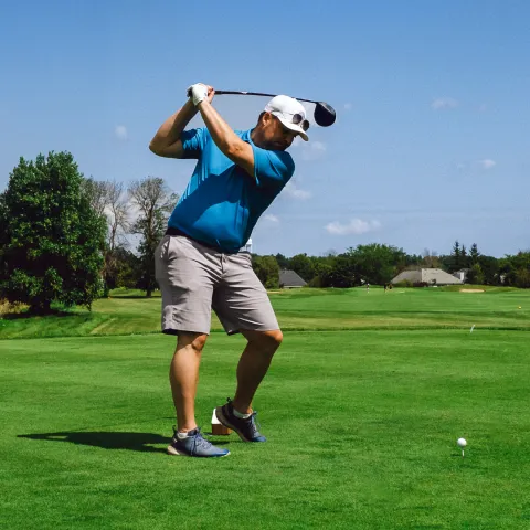 Man in blue polo shirt and beige shorts swinging a golf club.