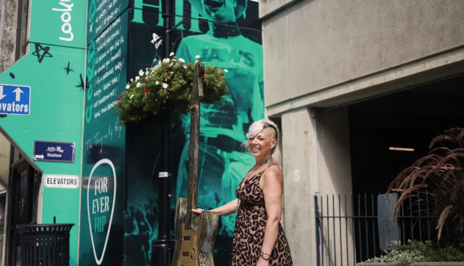 Cher Reynolds standing outside Hotel Dieu Hospital with her guitar art installation. The guitar stands at six feet tall and incorporates EGK lines in the design, weaving in the themes of healthcare to the Kingston story of music and art. In the background you see a wall wrap promoting The Tragically Hip's ten year anniversary since their last show.