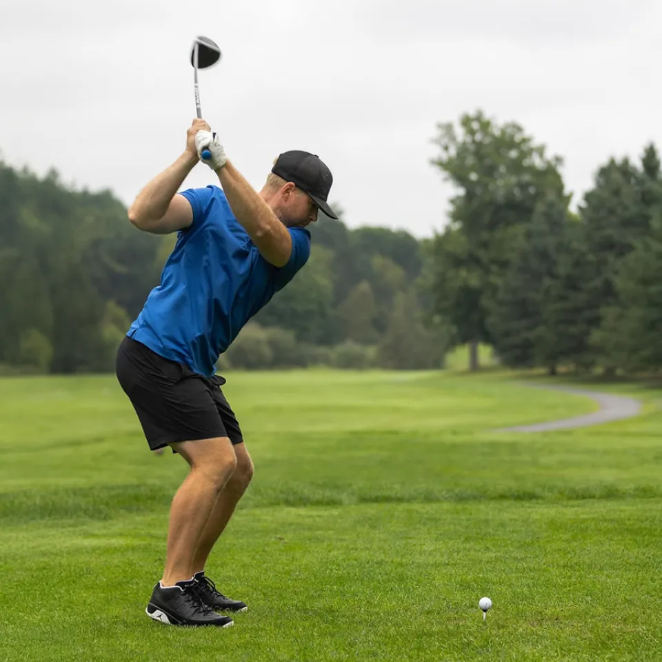 Man swinging golf club on the green in a blue golf shirt and black hat.